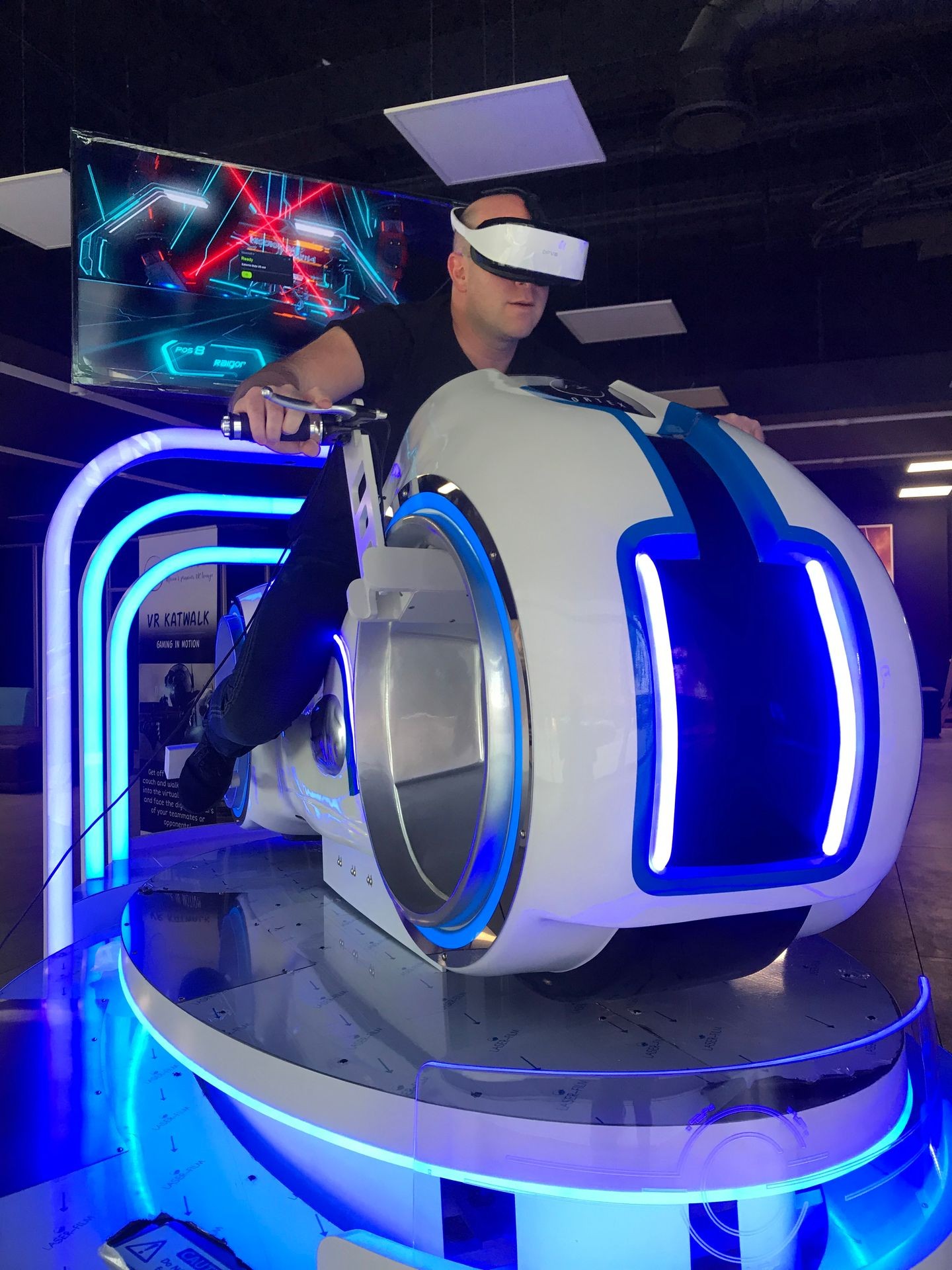 Person using a futuristic VR motorcycle simulator with glowing blue lights in an arcade setting.