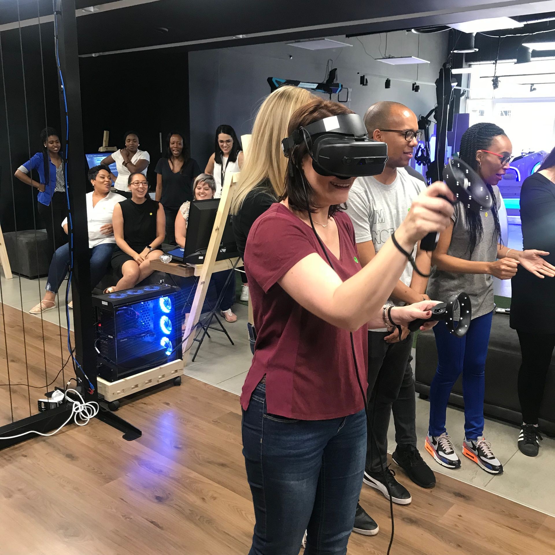 Person wearing a virtual reality headset and holding controllers in a room with several people and computer equipment.